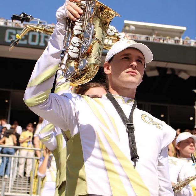 Close up of Hunter Ashcraft holding up a saxophone looking into the distance wearing a Georgia Tech marching band uniform. In the background is a stadium and other band members.